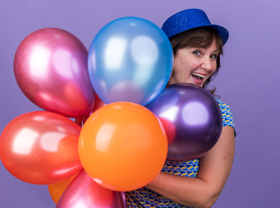 庆祝快乐而兴奋的中年妇女戴着派对帽 手里拿着一束五颜六色的气球 微笑着站在紫色的墙上庆祝生日派对微笑束生日