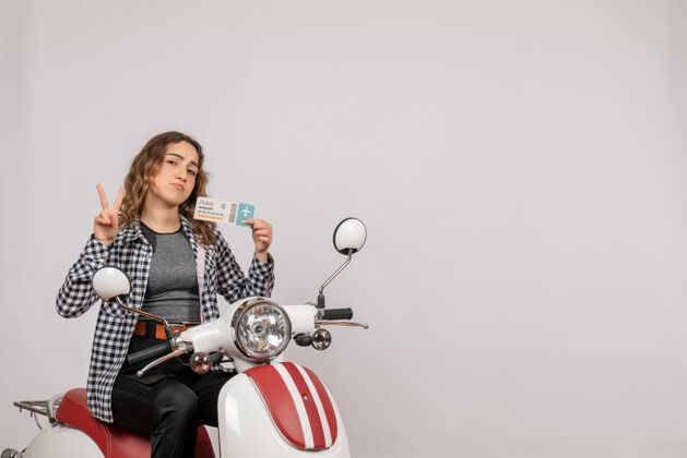肖像骑着轻便摩托车的年轻漂亮姑娘 手里拿着一张票 在灰色地上做着一个好牌子摩托车时尚灰色