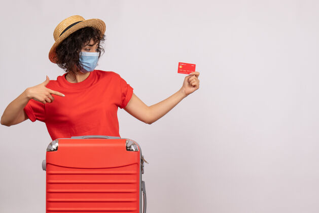 持有正面图：年轻女性 戴着面具 手里拿着白色背景上的红色银行卡 太阳光照射着大流行的度假旅游彩钱旅行银行面具