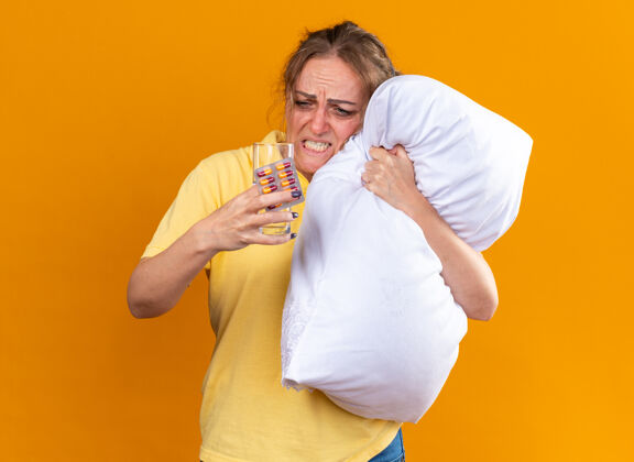 女人不健康的女人穿着黄色衬衫 患流感和感冒 感觉不舒服 抱着枕头 拿着药片和一杯水 站在橘色的墙上 看起来很烦躁和失望不适枕头站着