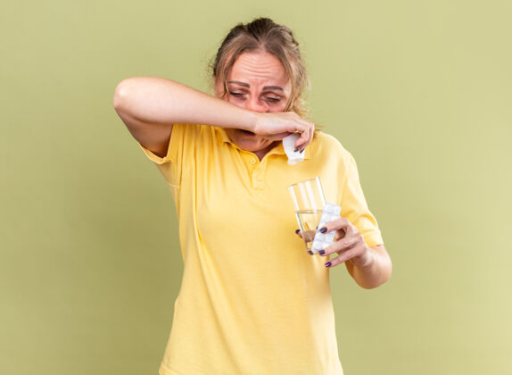 抱着一个穿着黄色衬衫的不健康的女人站在绿色的墙壁上 手里拿着一杯水和药片擦着流鼻涕 感觉很糟糕站着不健康药片