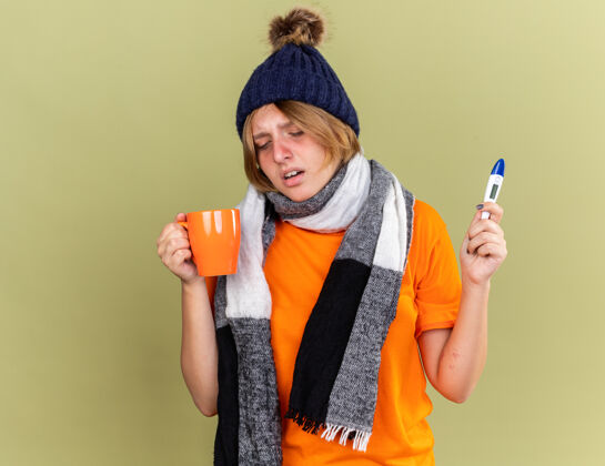 周围不健康的年轻女子戴着帽子 脖子上围着围巾 感觉不舒服 喝着热茶 拿着患流感的数字温度计站在绿色的墙上感觉穿着脖子
