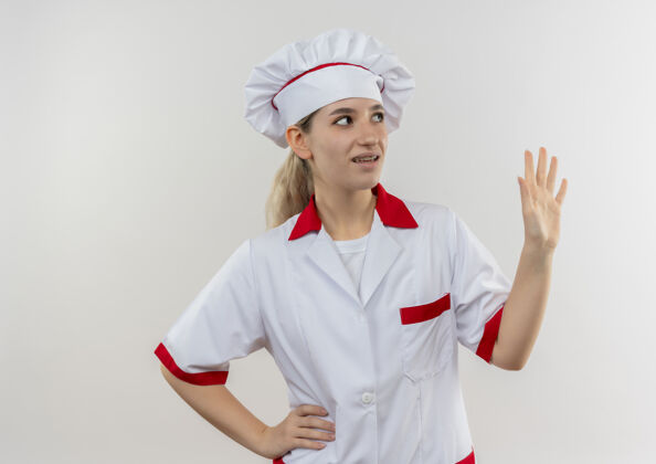 壁板年轻漂亮的厨师穿着厨师制服 戴着牙套 手放在腰上 举起手 看着隔离在白墙上的一边腰牙科制服