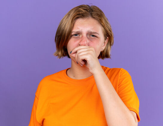 感觉不健康的年轻女子穿着橙色t恤 感觉很糟糕擦鼻子发烧咳嗽感冒站在紫色的墙上年轻不健康鼻子