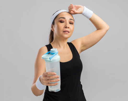 站着戴着头带和臂章拿着水瓶站在白墙上看起来很累的年轻健身女士臂章奶瓶头带