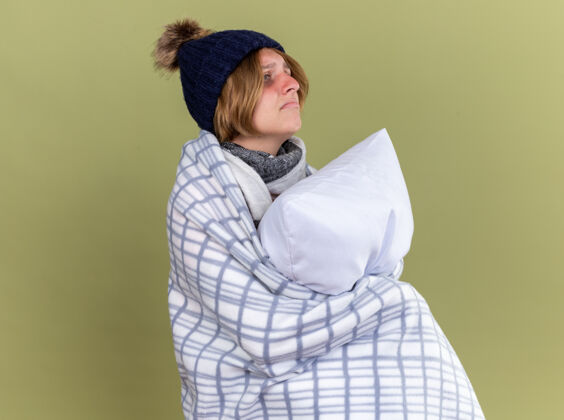 表情不健康的年轻女子裹着毛毯戴着帽子抱着枕头患流感感觉不舒服站在绿色的墙边带着悲伤的表情看着一边毯子裹着抱着