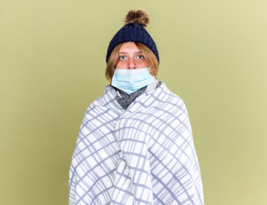 温暖不健康的年轻女子戴着暖和的帽子 裹着毛毯 戴着面罩 站在绿色的墙上感觉不舒服 患流感不健康女人面具