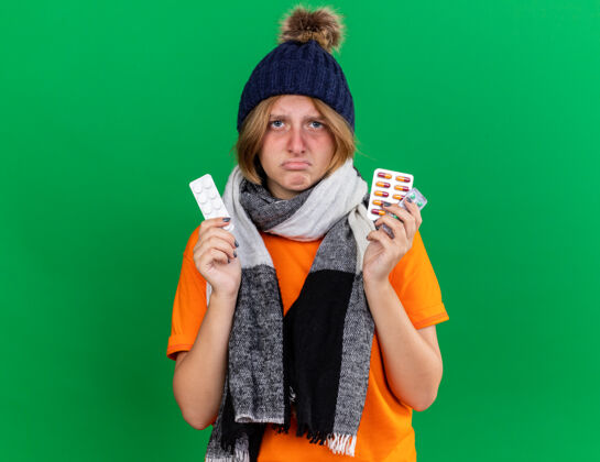 站不健康的年轻女子穿着橙色t恤 戴着帽子 脖子上围着暖和的围巾 感觉很可怕 拿着病毒的药片站在绿色的墙上抱温暖周围