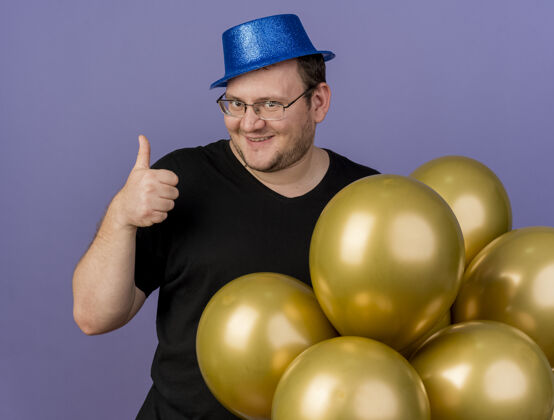 蓝色令人印象深刻的成年斯拉夫人戴着眼镜戴着蓝色派对帽竖起大拇指站在氦气球氦聚会气球