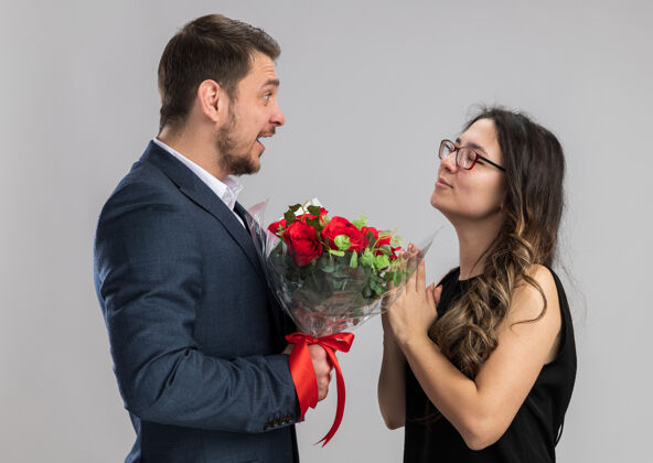 请一对年轻漂亮的情侣手拿着一束玫瑰看着他幸福快乐的女友幸福地恋爱在白色的墙上庆祝情人节年轻礼物情侣