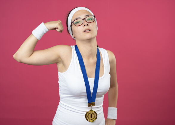 复制自信的年轻运动女孩戴着眼镜 脖子上戴着金牌 戴着头带和腕带 紧张的二头肌金色眼镜年轻
