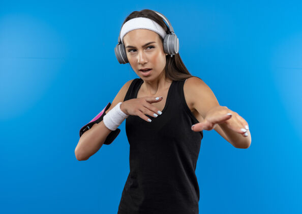 手自信的年轻漂亮的运动女孩戴着头带和腕带 戴着耳机 手上戴着电话袖标 望着蓝色墙上孤立的一面穿耳机运动
