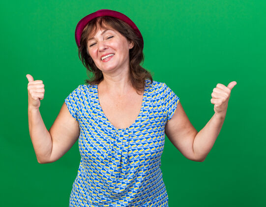 年龄戴着派对帽的中年妇女快乐快乐的微笑着竖起大拇指庆祝生日派对站在绿色的墙上站立女人生日