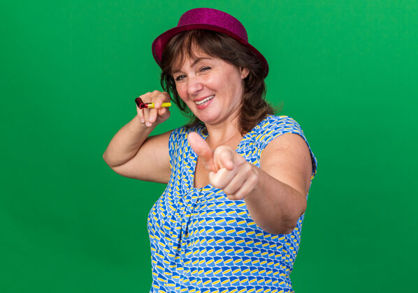 手指戴着派对帽的中年妇女手持哨子 食指微笑 站在绿色的墙上欢庆生日派对目录生日年龄