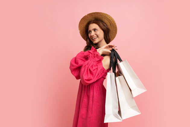 太阳镜时髦的红发女人 带着白色购物袋 穿着粉色连身裙 袖子搭在粉色的墙上春天高兴人