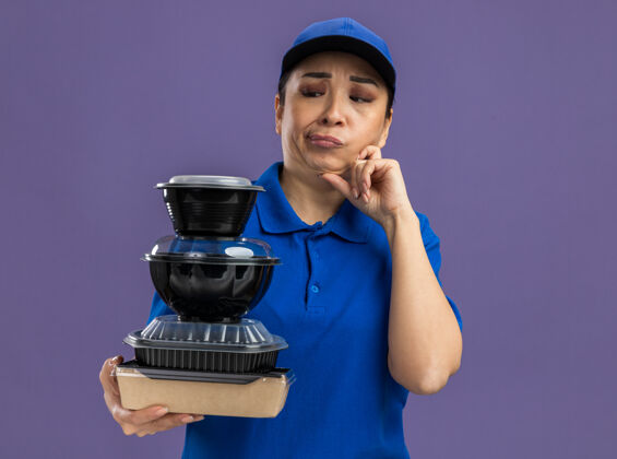 盒子身穿蓝色制服 戴着帽子的年轻送货员站在紫色的墙上 手里拿着一叠食品包 困惑地看着它们拿着制服送货