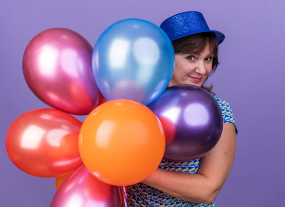 庆祝快乐开朗的中年妇女戴着派对帽手持一束五颜六色的气球微笑着站在紫色的墙上庆祝生日派对女人立场中间