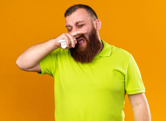 站着穿着黄色马球衫 留着不健康胡须的男人 站在橘色的墙上擦着流鼻涕 感觉很难受跑步鼻子感冒