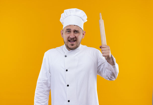 别针年轻帅气的厨师穿着厨师制服拿着擀面杖孤立地站在橙色的墙上帅气卷厨师