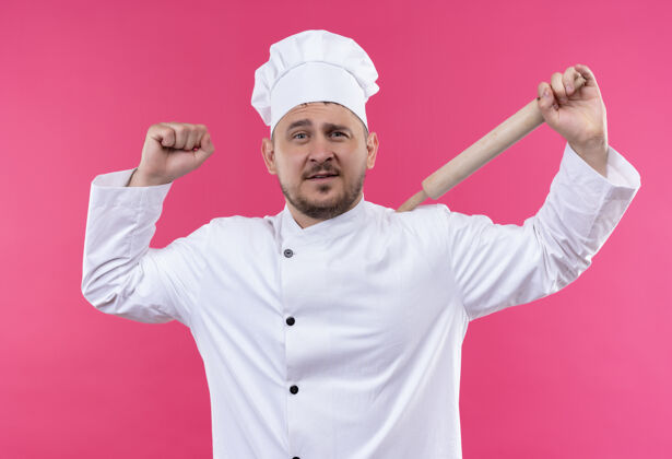 烹饪自信的年轻帅哥厨师穿着厨师制服拿着擀面杖在粉红色的墙上做着坚强孤立的手势强壮别针帅气