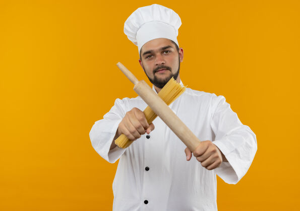 男性自信的年轻男性厨师身着厨师制服 将擀面杖和意大利面伸向隔离的橙色墙壁 留有复制空间年轻滚动伸展