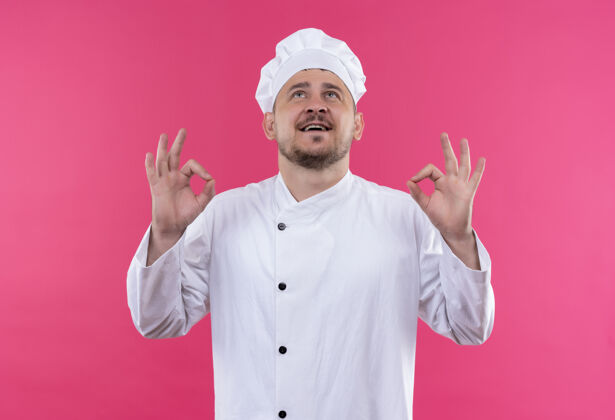 年轻快乐的年轻帅哥厨师身着厨师制服 抬头看着粉红色墙上的“做得好”标志制服厨师好的