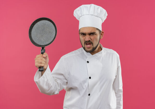 烹饪愤怒的年轻男厨师身着厨师制服 手拿煎锅孤立地站在粉红色的墙上厨师愤怒薯条
