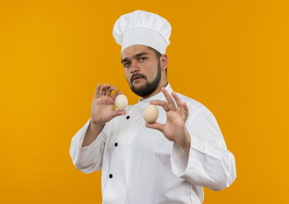 信心自信的年轻男性厨师身着厨师制服 手里拿着隔离在橙色墙上的鸡蛋烹饪年轻厨师