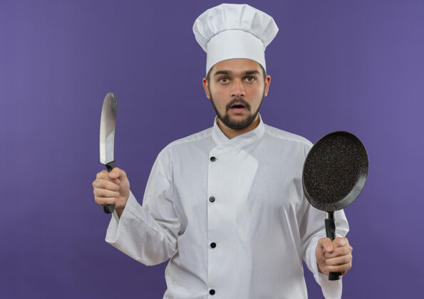 薯条穿着厨师制服的年轻男厨师手拿菜刀和煎锅孤立地站在紫色的墙上制服厨师印象