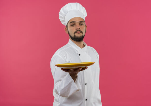 年轻自信的年轻男厨师身着厨师制服 将盘子伸向隔离的粉色墙壁 并留有复制空间烹饪自信厨师