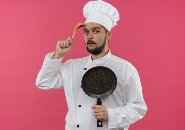 薯条穿着厨师制服的年轻男厨师手拿煎锅 用隔离在粉红色墙上的勺子摸头烹饪平底锅勺子