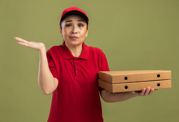 胳膊年轻的送货员身穿红色制服 头戴鸭舌帽 手举披萨盒 困惑地站在绿色的墙上女人盒子披萨