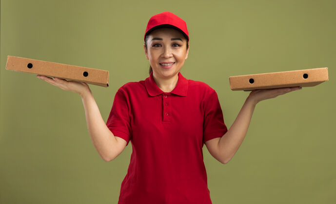 微笑身穿红色制服 头戴鸭舌帽 手拿披萨盒 友好微笑的年轻送货员站在绿色的墙上送货朋友站着