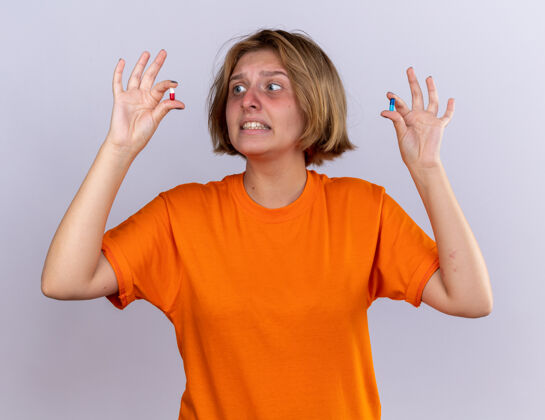 不健康不健康的年轻女子穿着橙色t恤 感觉身体不适 手上拿着感冒药 站在白色的墙上显得困惑和担心女人吃药迷茫
