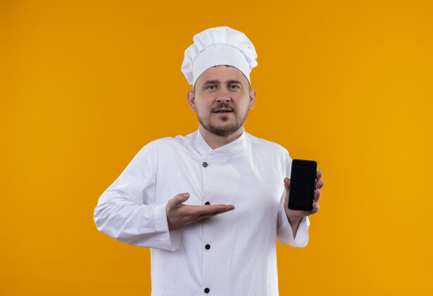 帅气自信的年轻帅哥厨师身着厨师制服 手持手机 孤立地指着橙色墙上的手机制服烹饪厨师