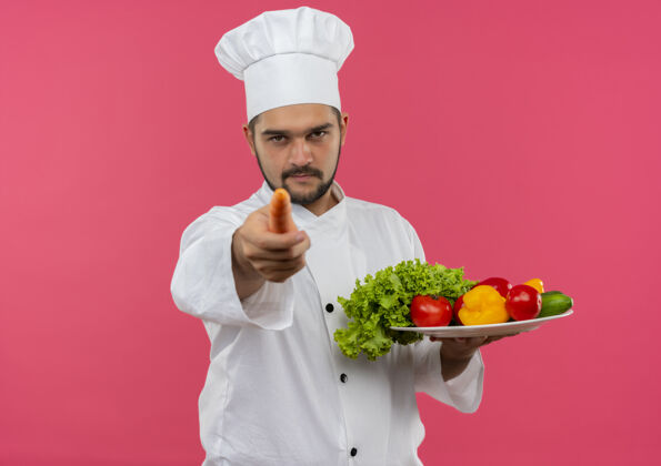 蔬菜自信的年轻男性厨师 身着厨师制服 端着一盘蔬菜 用胡萝卜指着粉色墙壁 并留有复制空间烹饪指向男性