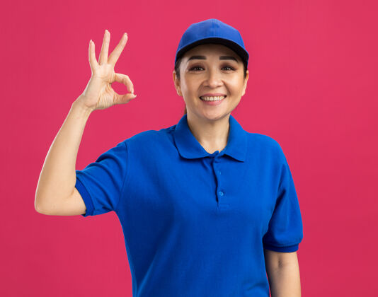 好身穿蓝色制服 头戴鸭舌帽的年轻女送货员站在粉红色的墙上高高兴兴地微笑着做“ok”的牌子女人站着微笑