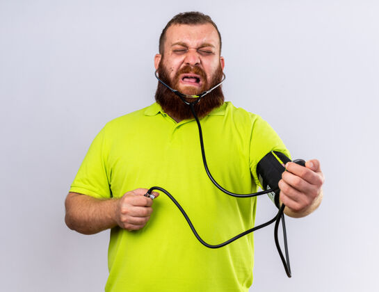 不健康不健康的胡子男人穿着黄色马球衫感觉不舒服用血压计测量血压站在白墙上看起来很沮丧测量压力男人