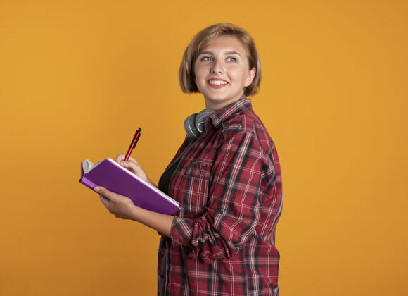 橙色微笑的年轻斯拉夫女学生戴着耳机 背着书包 站在一边拿着书和笔戴着年轻人耳机