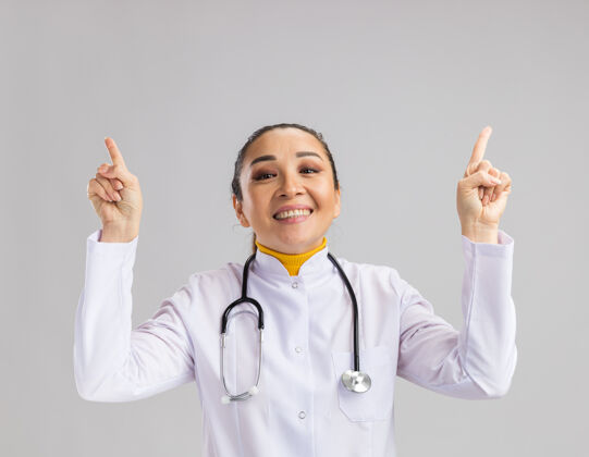 医生年轻的女医生穿着白色的医用外套 脖子上戴着听诊器 高兴地用食指站在白色的墙上站手指目录