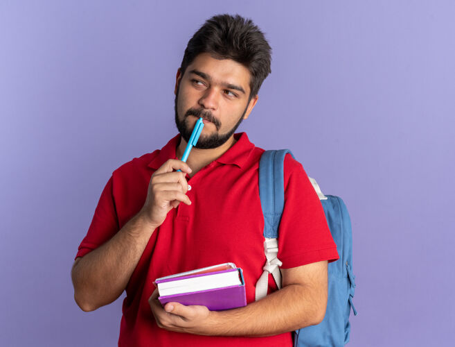 学生年轻的留着胡子的学生 穿着红色马球衫 背着书包 手里拿着书和笔 站在一旁困惑地看着年轻人马球靠边
