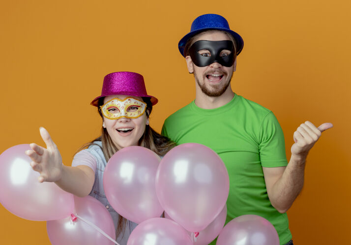 帅气快乐的年轻夫妇站在氦气球女孩在粉红色的帽子戴化装眼罩点向前与手帅哥在蓝色的帽子戴化装眼罩点在一边帽子眼睛侧线