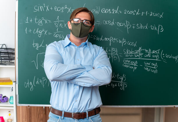 靠近戴着防护面具戴着眼镜的年轻男老师站在黑板旁 手里拿着数学公式 在教室里双臂交叉 看上去很自信面部男人站立