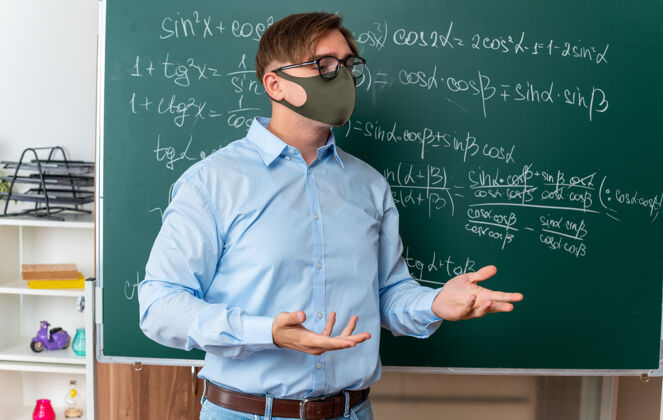 课戴着口罩戴着眼镜的年轻男教师站在黑板旁 用数学公式讲解课文 在教室里显得很困惑面部新常态佩戴