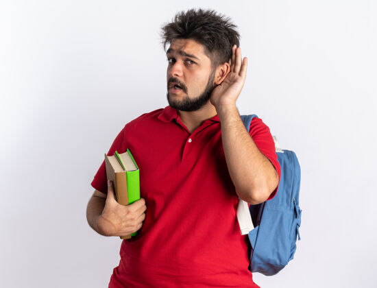 耳朵年轻的留着胡子的学生 穿着红色马球衫 背着背包 拿着笔记本 手放在耳朵上 站着试着听试试笔记本学生