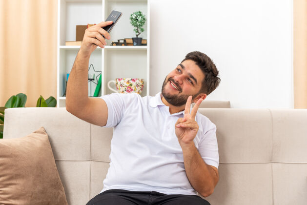 休闲穿着休闲服的年轻人用智能手机自拍快乐而积极地展示着v型标志微笑坐在客厅的沙发上手机坐着衣服