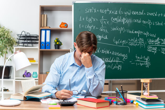 笔记戴着眼镜的年轻男老师累了 过度劳累了 闭着眼睛摸着鼻子坐在课桌旁 教室里黑板前放着书和笔记黑板男数学