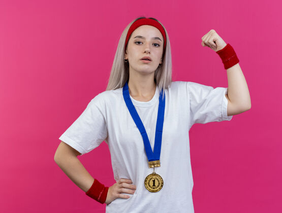 年轻自信的年轻白种人运动女孩 戴着牙套 脖子上戴着金牌 戴着头带和腕带 紧张的二头肌周围运动粉色