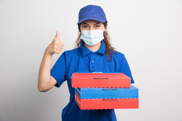 女性送比萨饼的女孩拿着三个盒子 戴着医用口罩 大拇指朝上竖着白色的帽子帽子手势