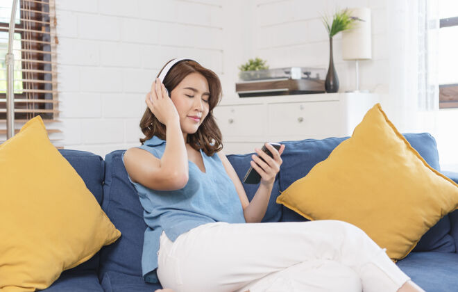 平静年轻的亚洲女性周末坐在客厅的沙发上 用智能手机和耳机听音乐音频床女士